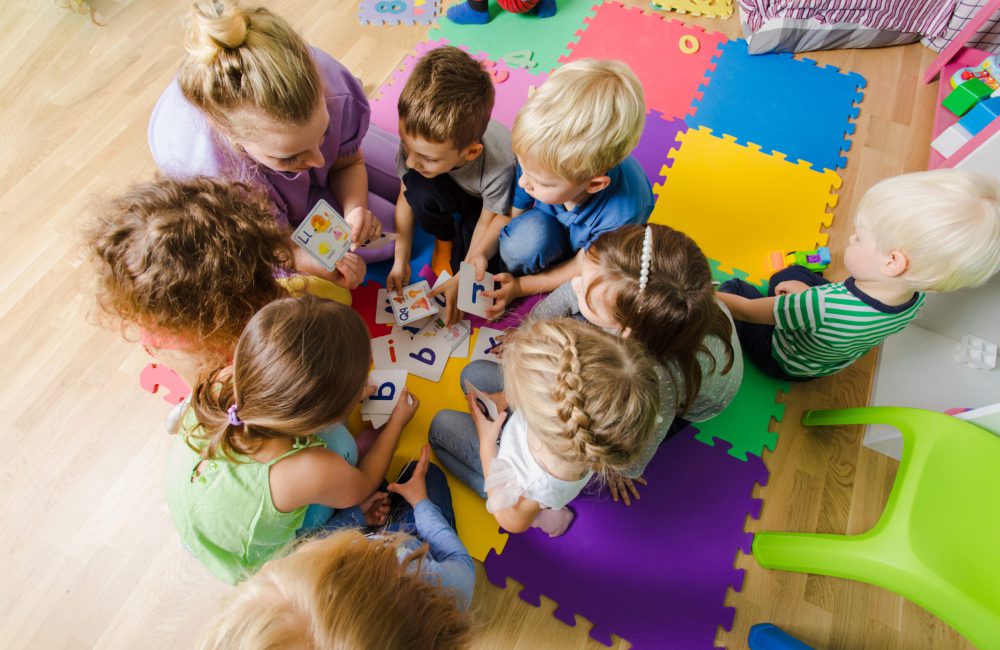 Group of kindergarten kids sitting closely on a floor together with teacher, providing group work. Children learning to cooperate while solving tasks.