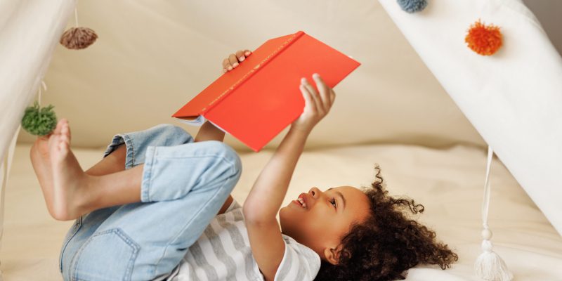 Portrait of cute little african american kid curly boy with book smiling at camera while   reading book  in play tent at home, happy child playing alone in children room